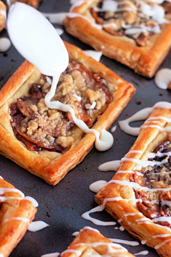 A close-up view of apple crumb danishes on a baking sheet, with a spoon drizzling white icing over one of the pastries.