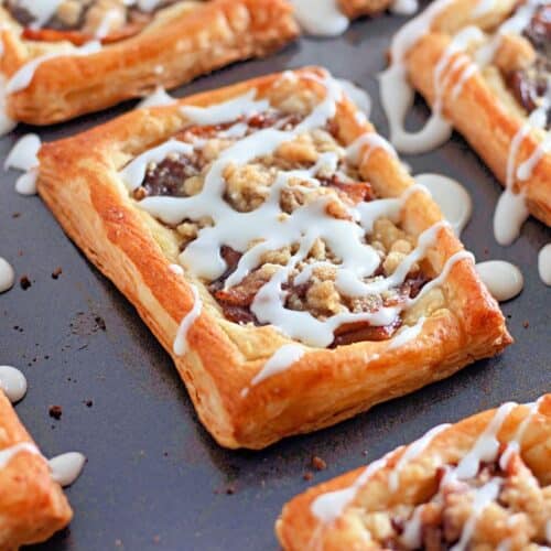 A close-up view of rectangular apple crumb danishes made with golden-brown puff pastry, drizzled with white icing and topped with a crumbly streusel.