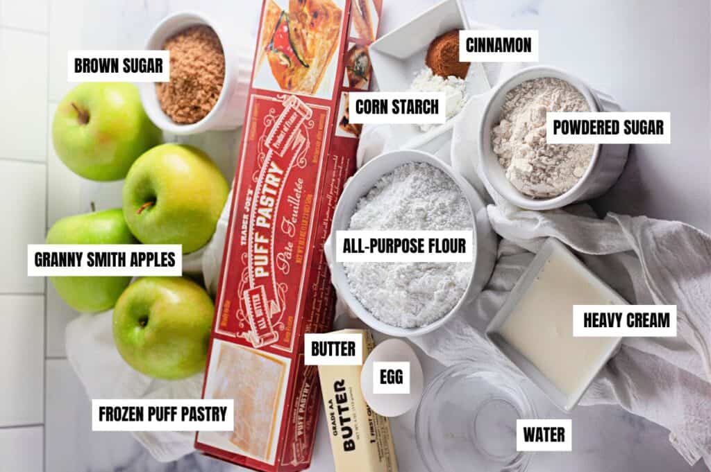 
Ingredients for apple crumb danishes arranged on a counter: Granny Smith apples, frozen puff pastry, brown sugar, cinnamon, corn starch, powdered sugar, all-purpose flour, butter, an egg, heavy cream, and a bowl of water, each labeled clearly.