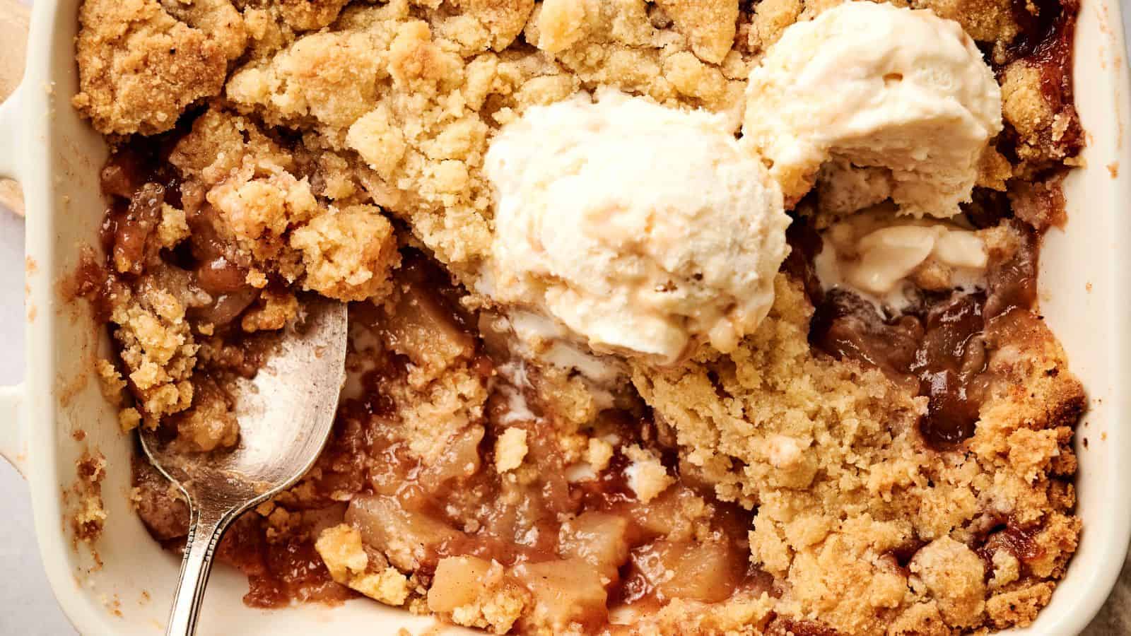 Close-up of an apple crumble dessert with a golden crust, topped with two scoops of vanilla ice cream, and a spoon placed inside the dish.