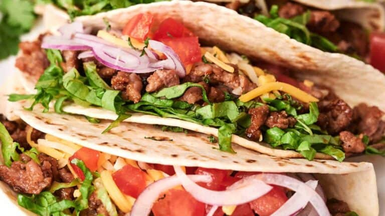 Three beef tacos filled with ground meat, shredded lettuce, diced tomatoes, grated cheese, and sliced red onions.