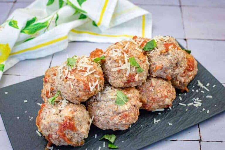 A stack of giant meatballs garnished with grated cheese and fresh herbs served on a black rectangular slate plate.