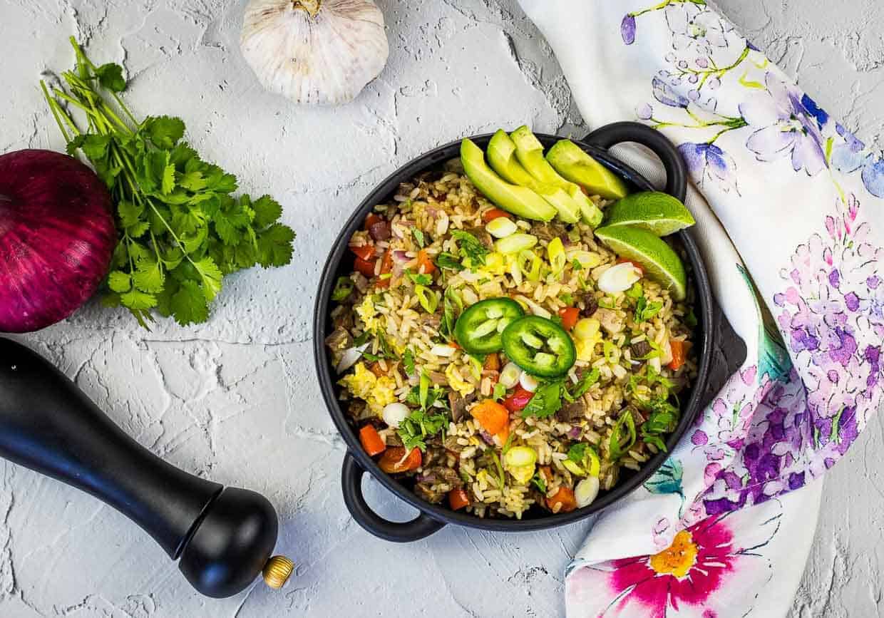 A cast-iron pan filled with Brisket Fried Rice, garnished with sliced avocado, lime wedges, jalapeño slices, and fresh cilantro.