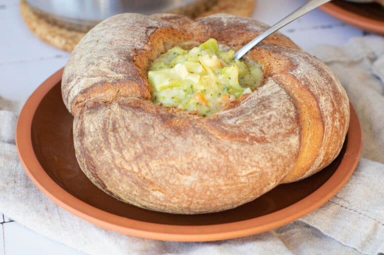 A hearty serving of creamy broccoli cheddar soup in a rustic sourdough bread bowl, with a spoon dipping into the soup, placed on a terracotta plate over a light linen cloth.