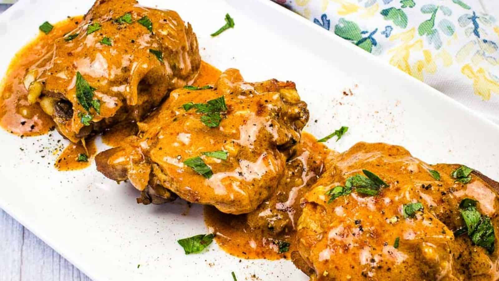 Three pieces of chicken smothered in a creamy, orange-hued sauce topped with chopped herbs, served on a white rectangular plate.