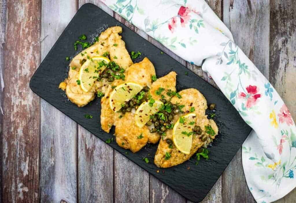 Chicken piccata on a black rectangular plate with parsley and lemon wedges.