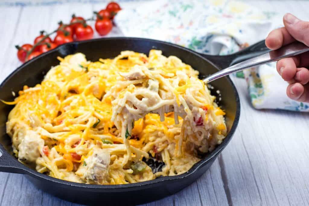 A cast-iron skillet filled with creamy chicken spaghetti, topped with melted cheddar cheese. A hand is lifting a portion with a fork, showing the cheesy, stringy texture.