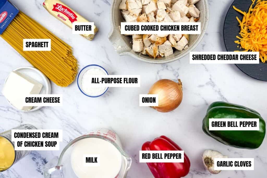 Ingredients for a chicken spaghetti recipe, including spaghetti, butter, cubed cooked chicken breast, shredded cheddar cheese, cream cheese, all-purpose flour, condensed cream of chicken soup, milk, onion, red bell pepper, green bell pepper, and garlic cloves. Items are arranged on a marble countertop.