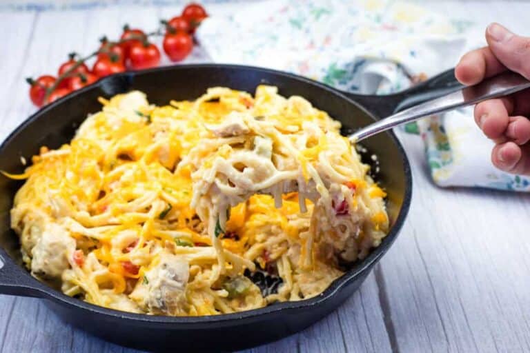 A hand holding a fork with a serving of chicken spaghetti from a cast-iron skillet.