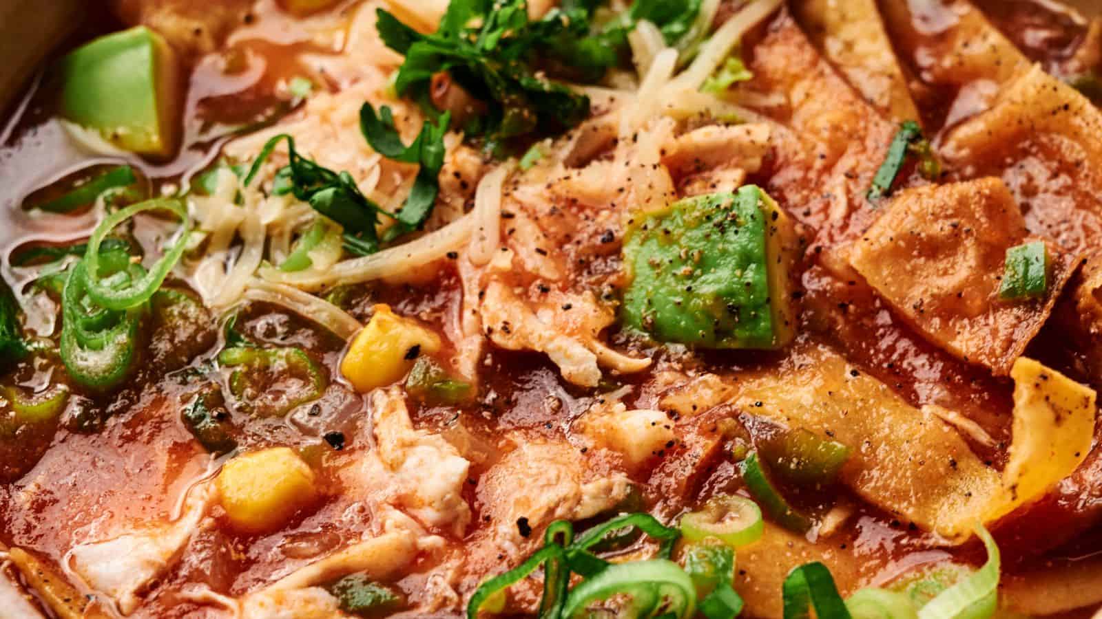 Close-up of a bowl of chicken tortilla soup with crispy tortilla strips, avocado pieces, corn, cheese, and herbs in a rich red broth.