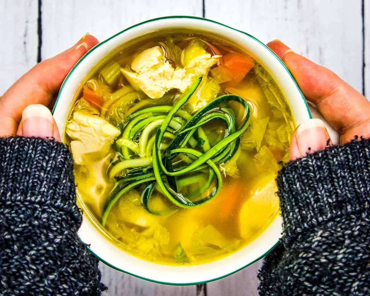 A person holding a bowl of chicken and zucchini soup.