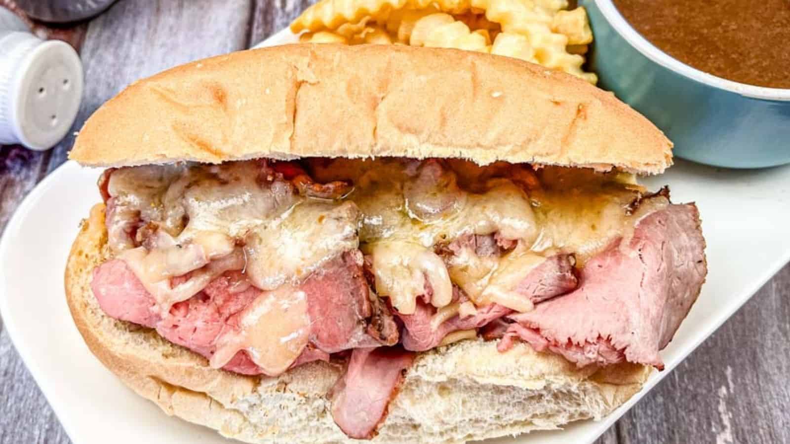 A roast beef sandwich with melted cheese on a hoagie roll, accompanied by crinkle-cut fries and a bowl of brown dipping sauce on the side.