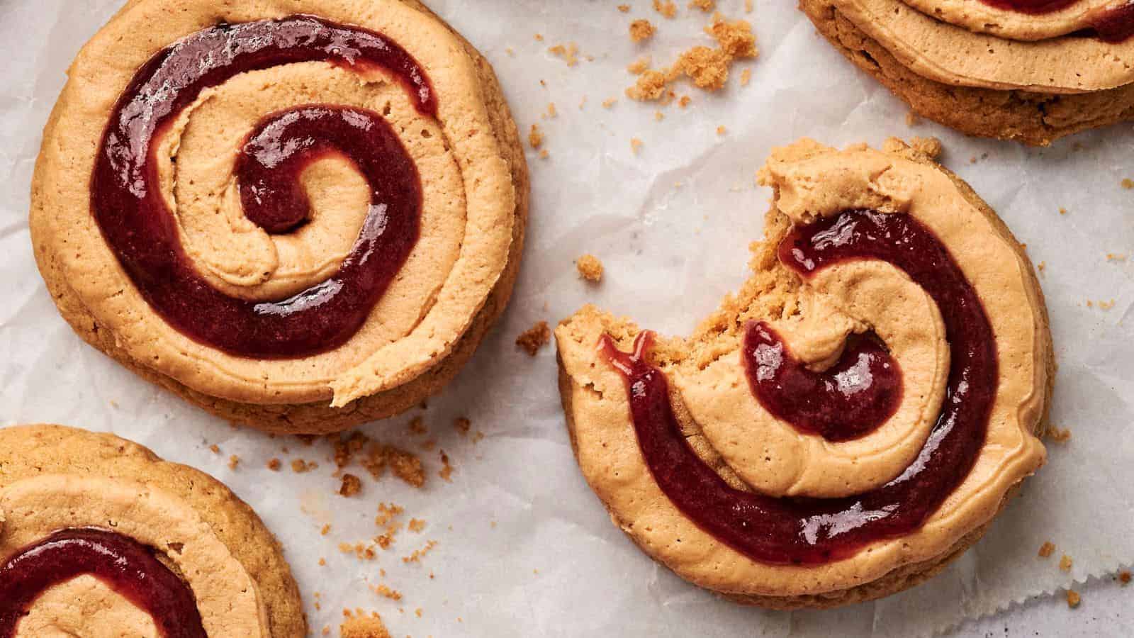 Close-up of spiral cookies with red jam filling on parchment paper, reminiscent of Copycat Crumbl Peanut Butter and Jelly Cookies, one partially eaten and surrounded by crumbs.