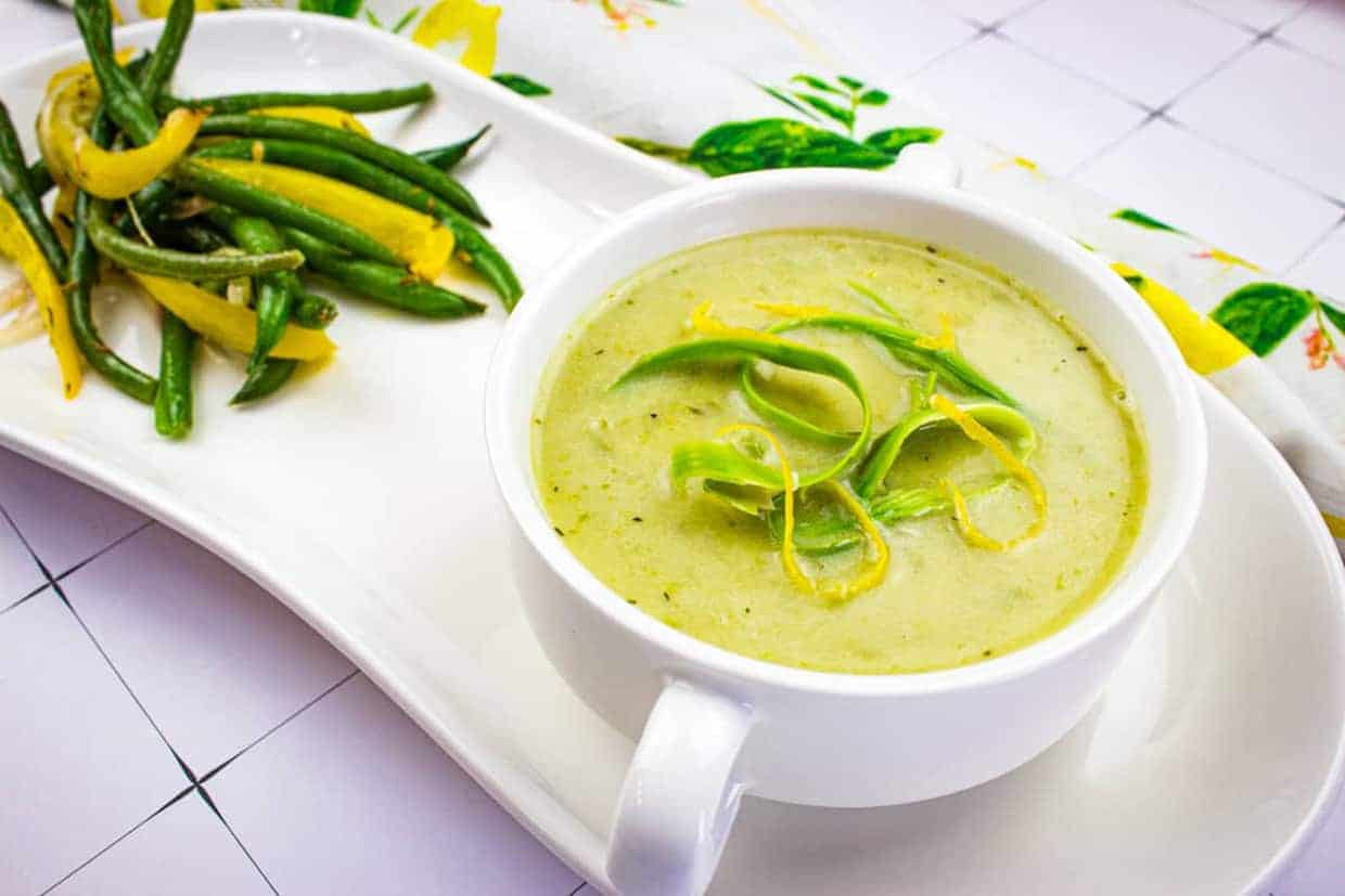 A bowl of Cream of Asparagus Soup garnished with asparagus ribbons and lemon zest, served alongside asparagus and lemon zest on a white platter.