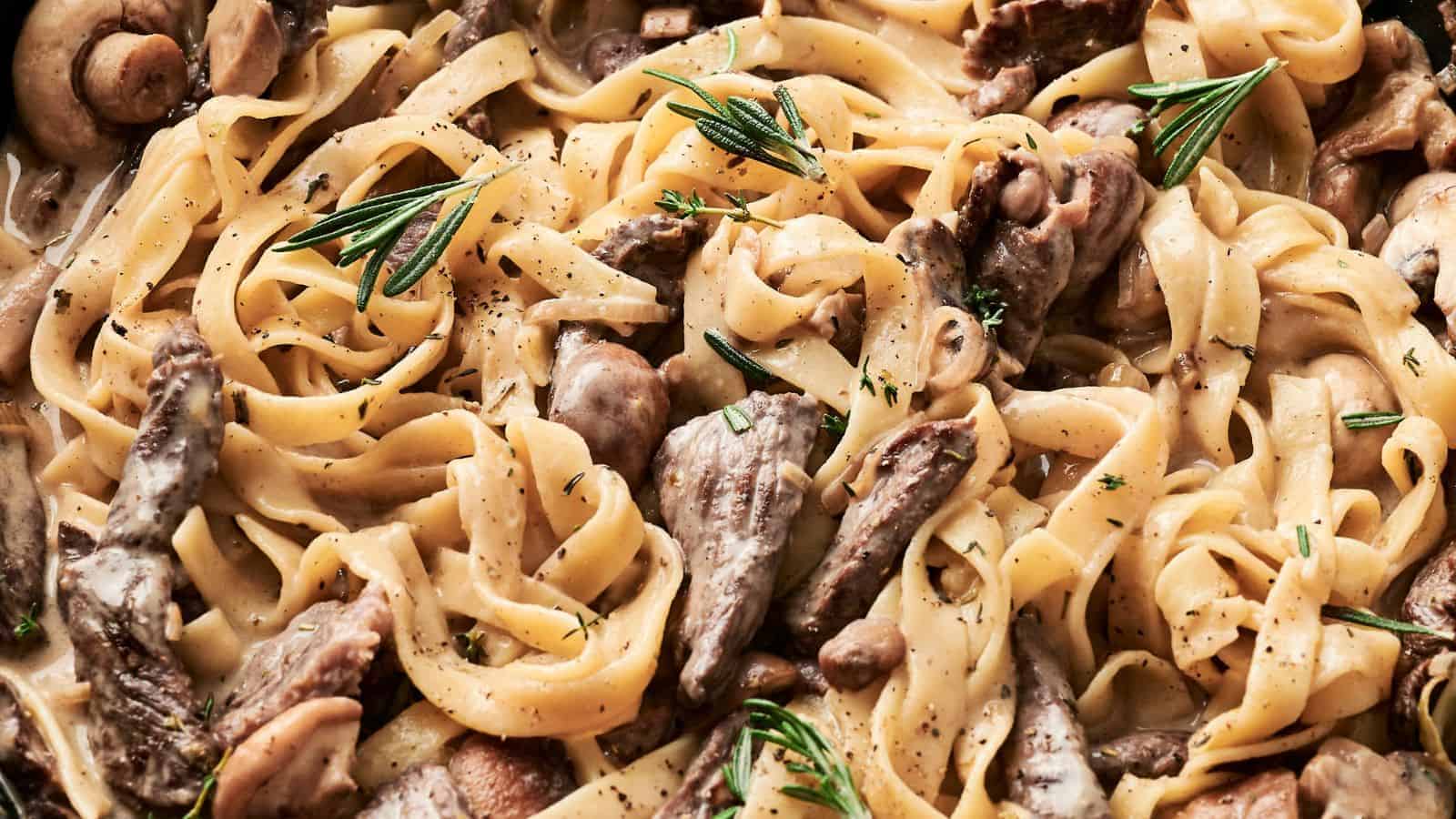 A close-up of fettuccine pasta with creamy sauce, reminiscent of Beef Stroganoff, paired with sliced beef, mushrooms, and sprigs of rosemary.