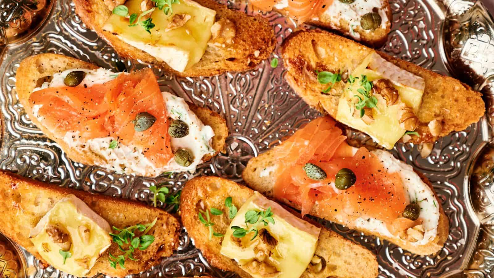 Toasted bread topped with smoked salmon, capers, cheese, walnuts, and herbs on a decorative silver tray.