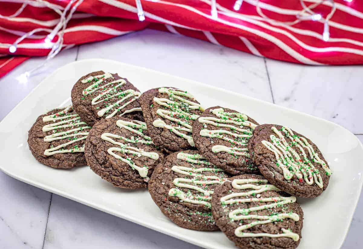 A plate of chocolate cookies topped with white icing and green sprinkles. A red and white striped cloth is in the background.