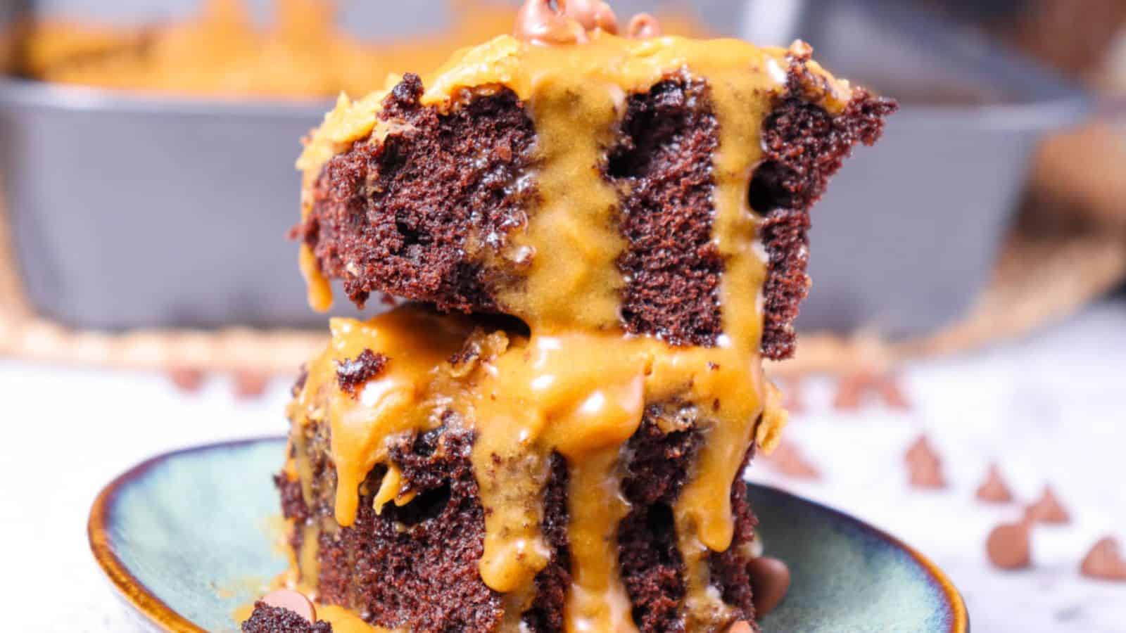 Close-up of two stacked pieces of chocolate cake drizzled with caramel sauce on a plate, with more cake in the background.