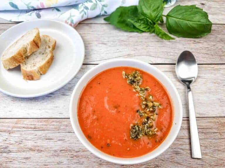 A bowl of Red Pepper and Tomato Soup with a spoon and bread on a plate on the side.