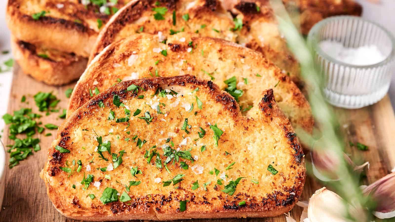 Slices of garlic bread topped with chopped parsley and sea salt sit enticingly on a wooden board, accompanied by a small bowl of salt and fresh cloves of garlic.