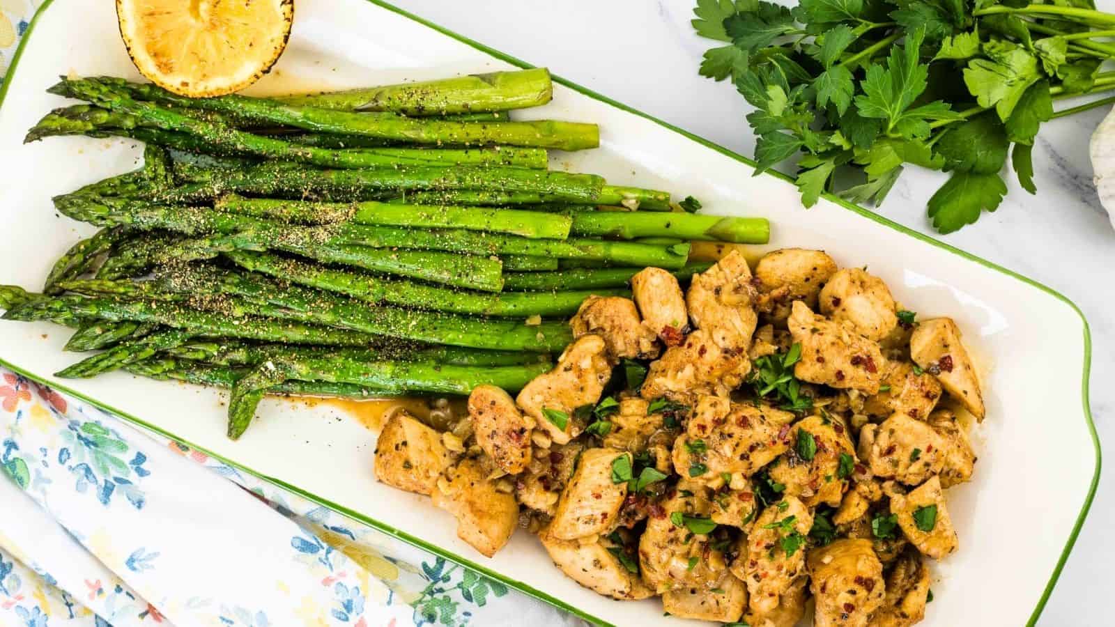 A plate with seasoned asparagus and garlic butter chicken bites topped with herbs. A halved grilled lemon and fresh parsley are nearby on a white floral tablecloth.