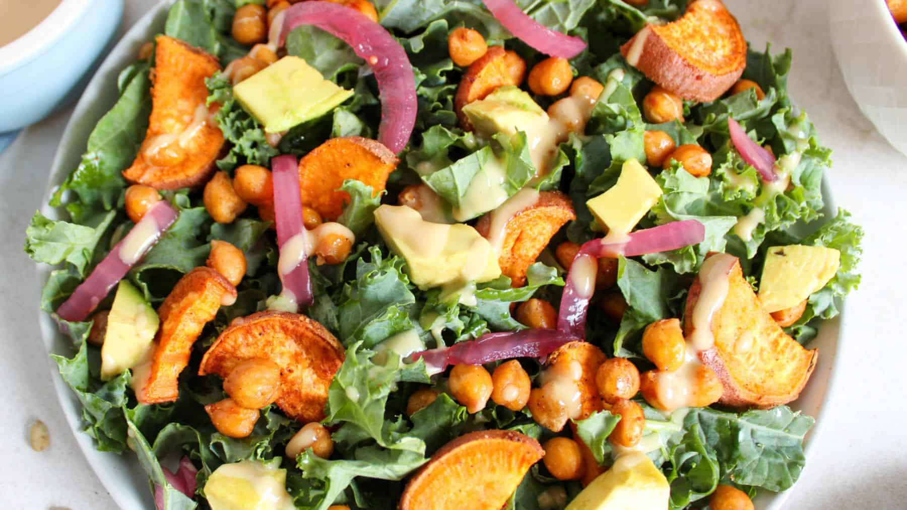 A Harvest Salad with kale, sweet potato wedges, avocado chunks, chickpeas, and pickled red onions, all drizzled with a delectable dressing.