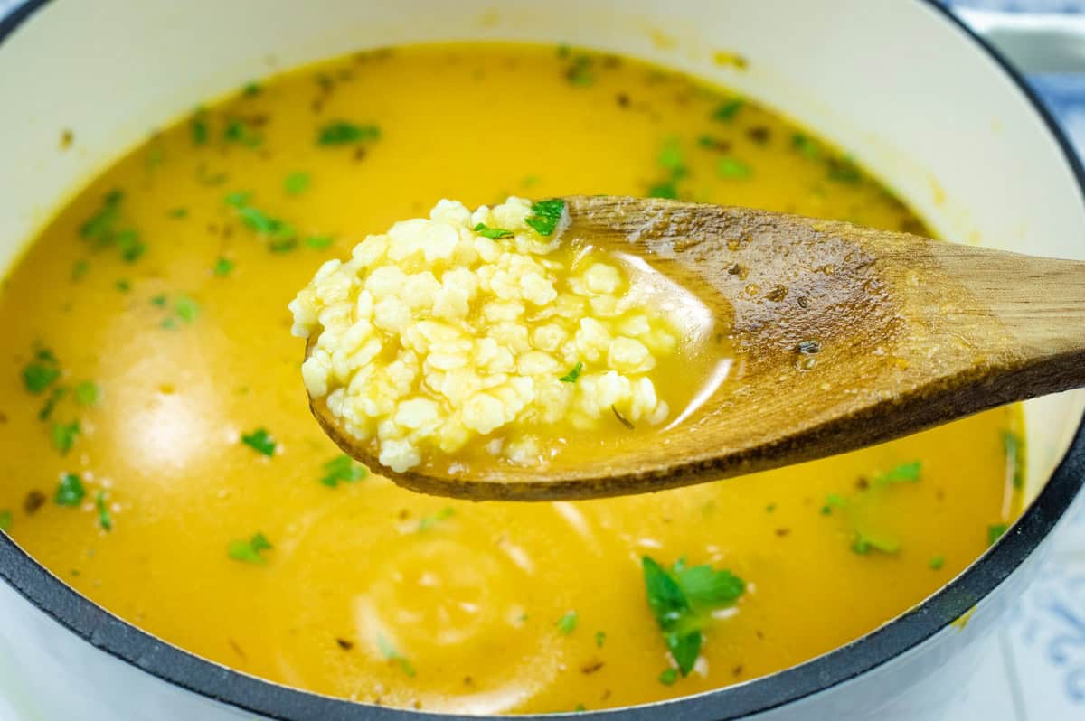 A wooden spoon holds a portion of Pastina Soup hovering above a pot.