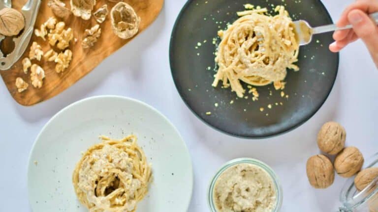 Italian walnut sauce pasta served on black and white plates, accompanied by a wooden board with shelled and unshelled walnuts, a jar of walnut sauce, and a fork twirling pasta on the side.