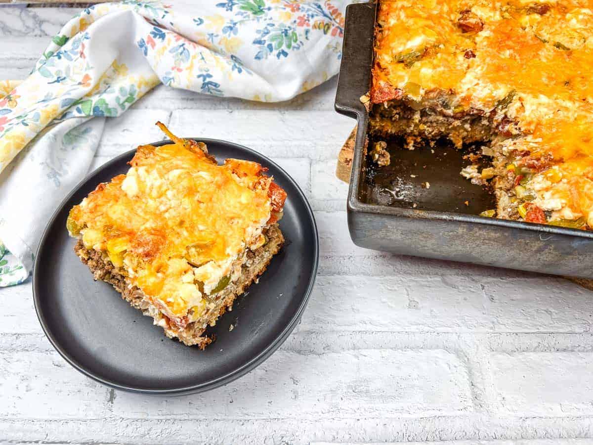 A slice of John Wayne casserole on a black plate beside a baking dish of the same casserole.