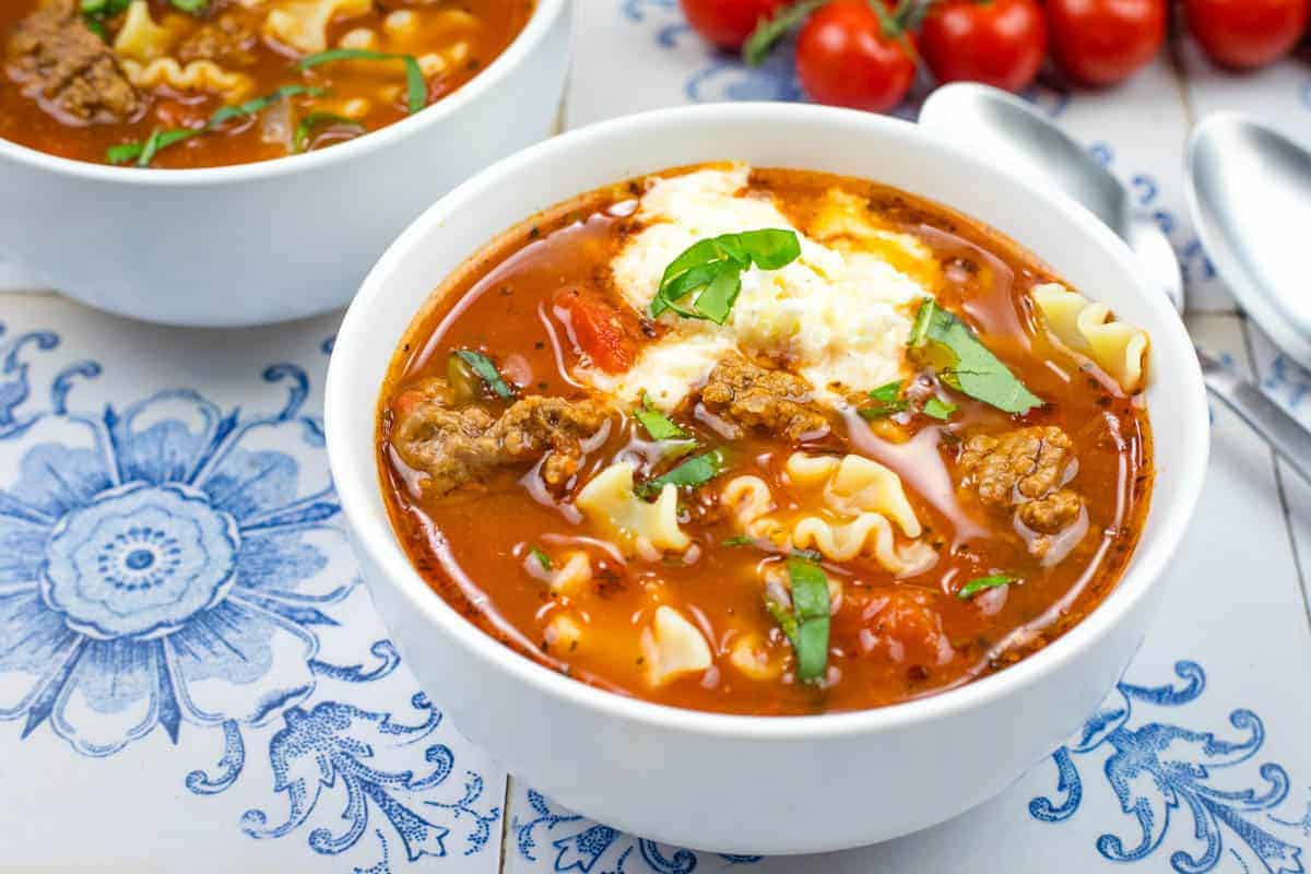 Close-up of a bowl of soup with lasagna soup, topped with chopped basil, on a blue-patterned tiles.