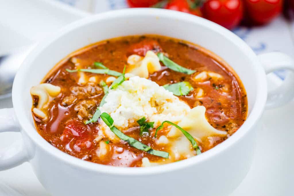 A bowl of beefy lasagna soup with a dollop of ricotta cheese, garnished with basil.