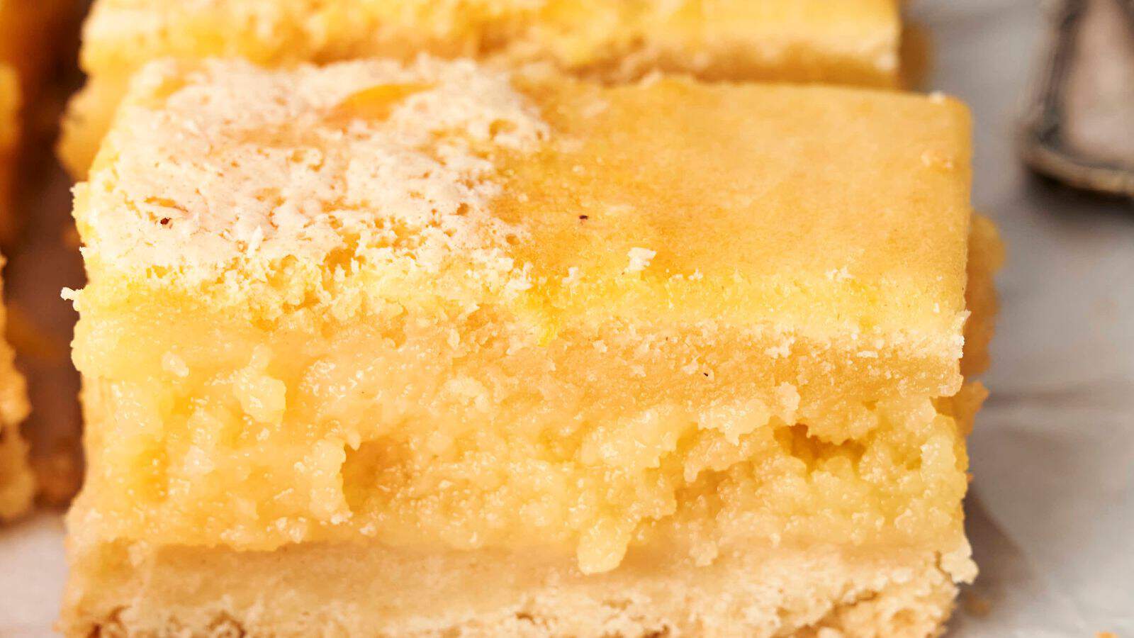 Close-up of a lemon bar with a crumbly, light yellow top layer and a thicker, moist filling resting on a shortbread crust.