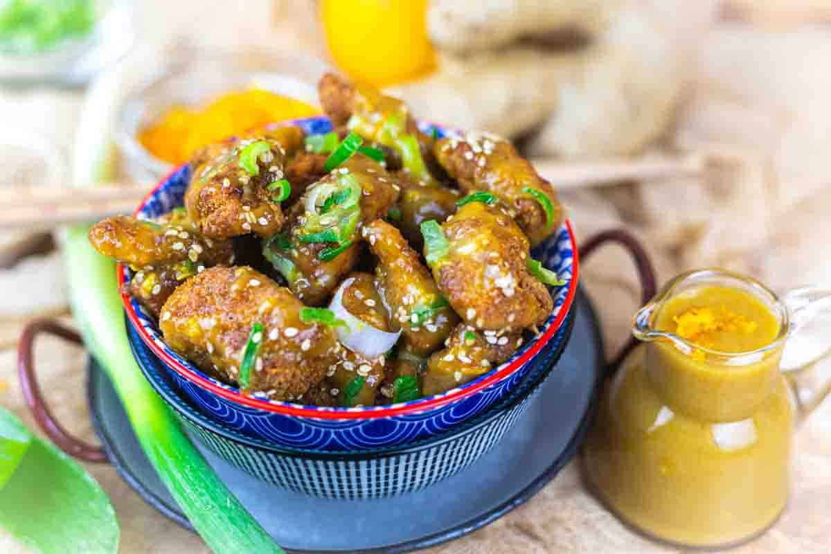 A bowl of fried chicken pieces coated in sauce and garnished with sesame seeds and green onions, accompanied by a small jug of sauce.