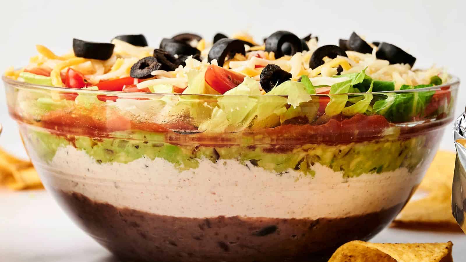 A clear glass bowl with 7 Layer Dip featuring beans, white cream, guacamole, diced tomatoes, shredded cheese, lettuce, and black olives.