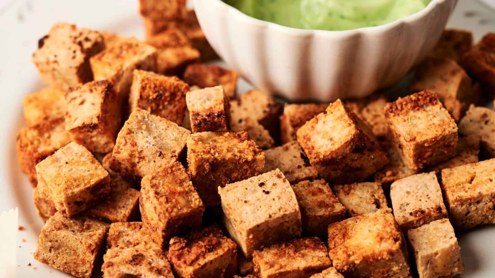 Crispy tofu cubes piled on a plate with a dish of green dipping sauce in the background.