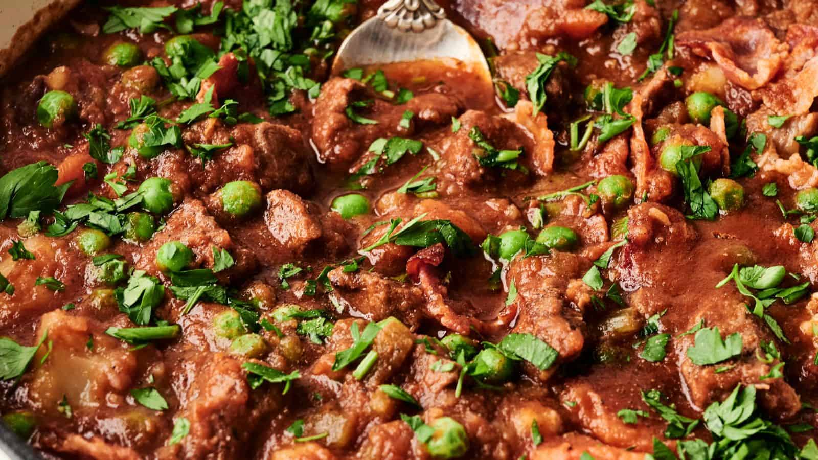 A close-up of a beef stew with green peas and chopped parsley, showing a spoon partially submerged in the rich, red sauce.