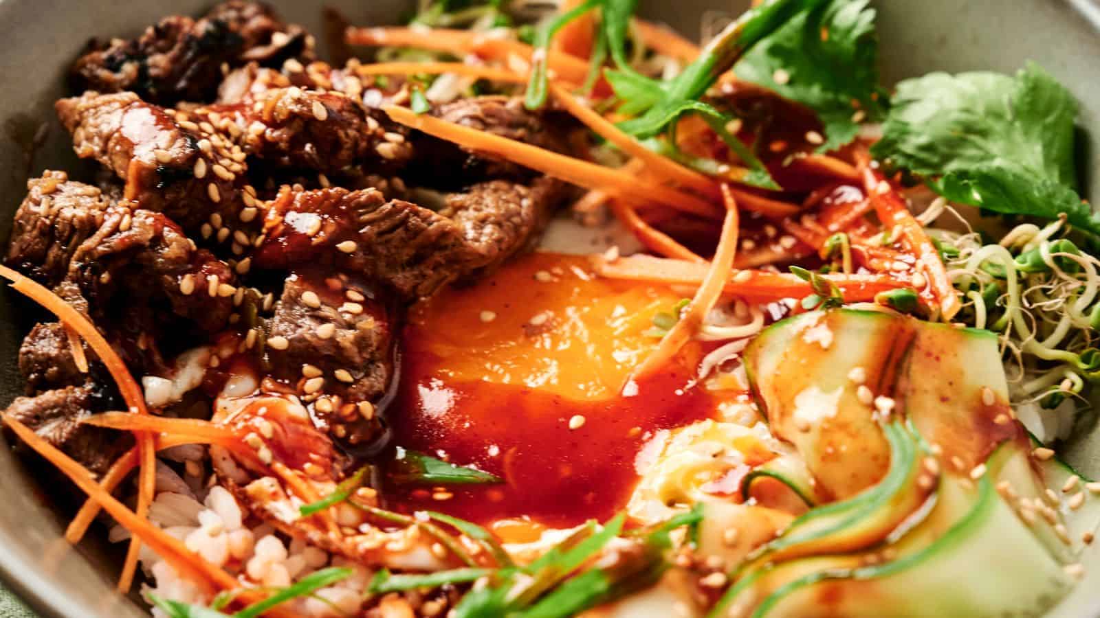 A bowl of bibimbap with sliced beef, egg yolk, carrots, cucumber, greens, and bean sprouts, topped with sesame seeds and red sauce.