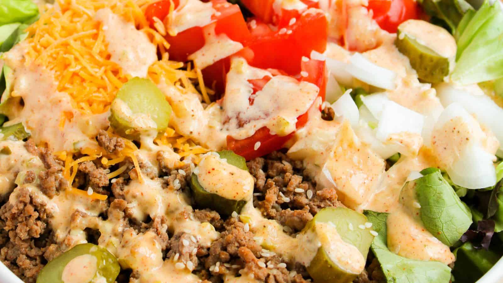 Close-up of a salad featuring ground beef, sliced pickles, diced tomatoes, shredded cheese, and diced onions, topped with a creamy dressing.