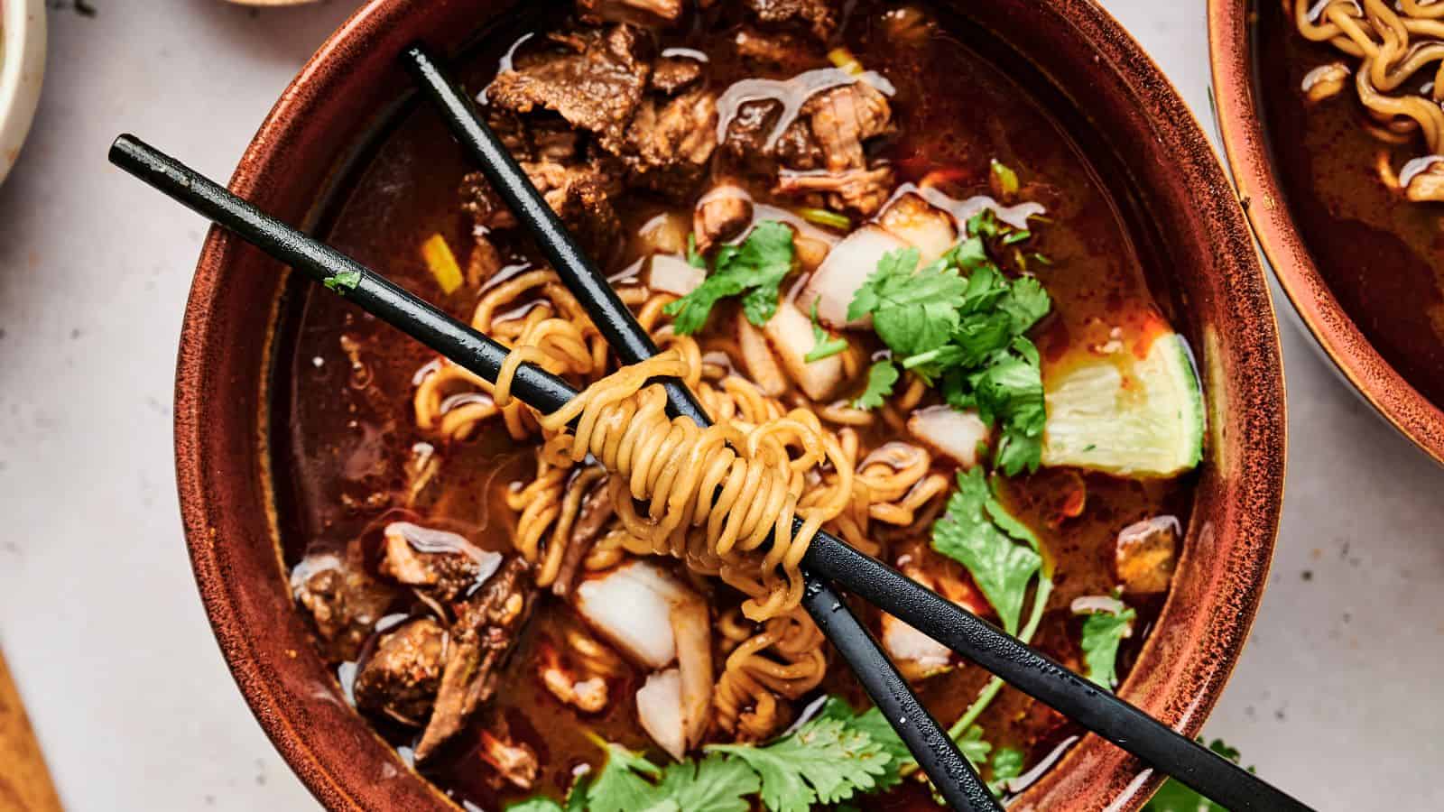 A bowl of ramen with meat, cilantro, and a lime wedge, topped with noodles wrapped around chopsticks.