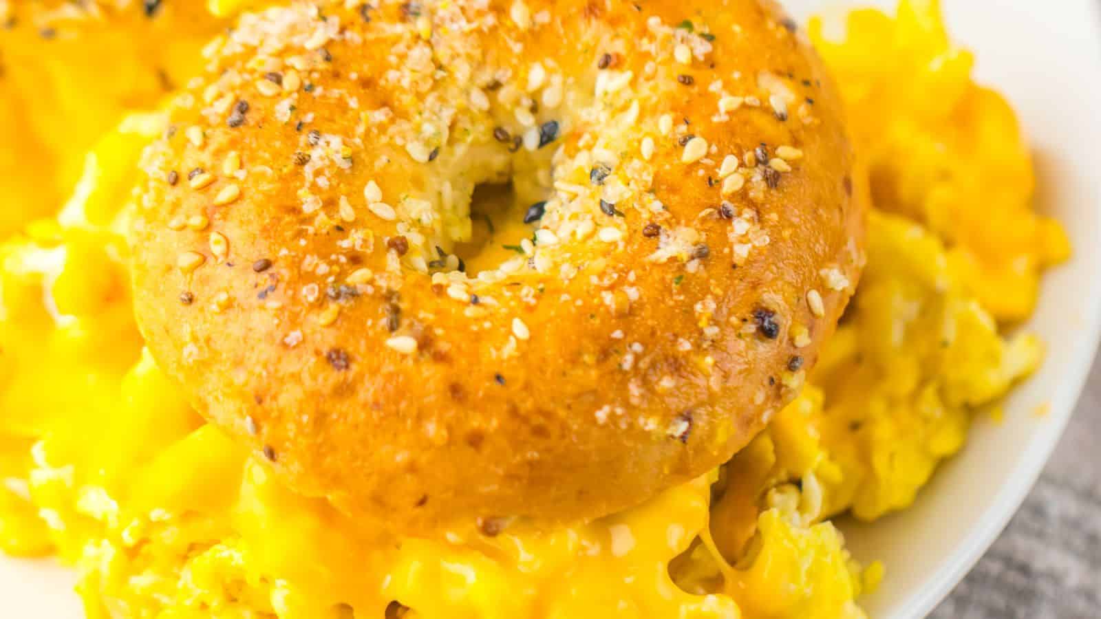 A sesame seed bagel sits on top of a serving of scrambled eggs with melted cheese on a white plate.