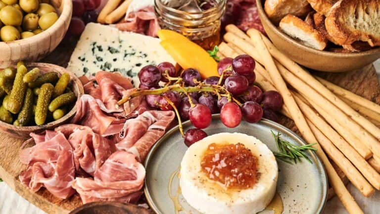 Charcuterie Board showcasing prosciutto, pickles, green olives, blue cheese, breadsticks, baguette slices, grapes, honey, and a cheese wheel topped with jam.