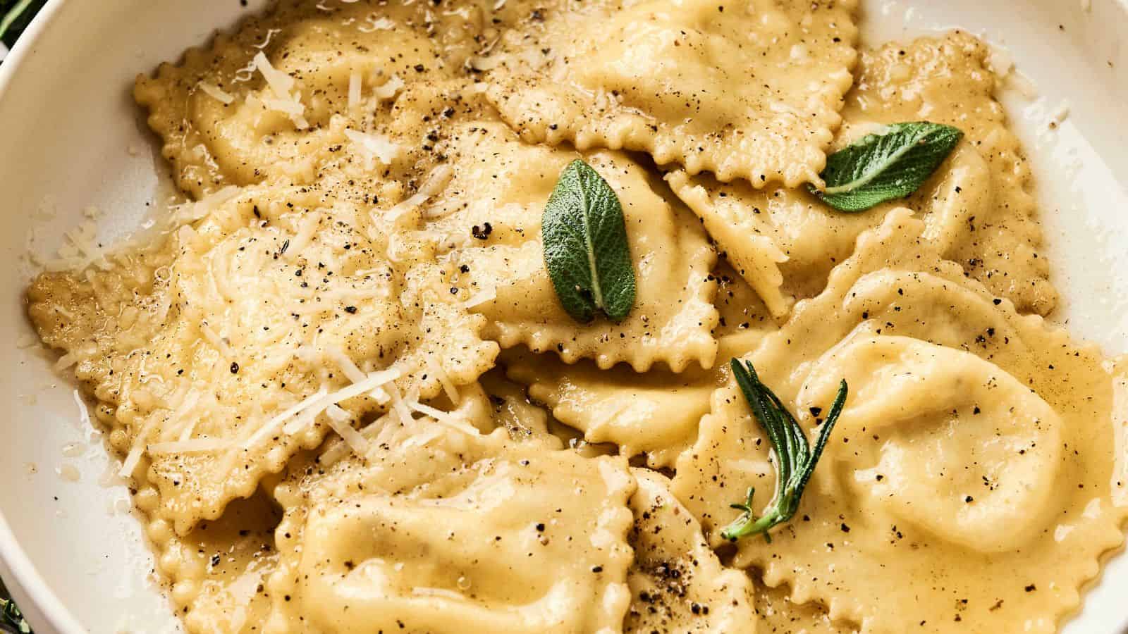 A white plate of ravioli topped with black pepper, grated cheese, and sage leaves.