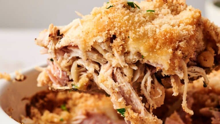 Close-up of a serving of baked chicken with a golden breadcrumb crust being scooped from a dish, revealing tender shredded meat underneath.