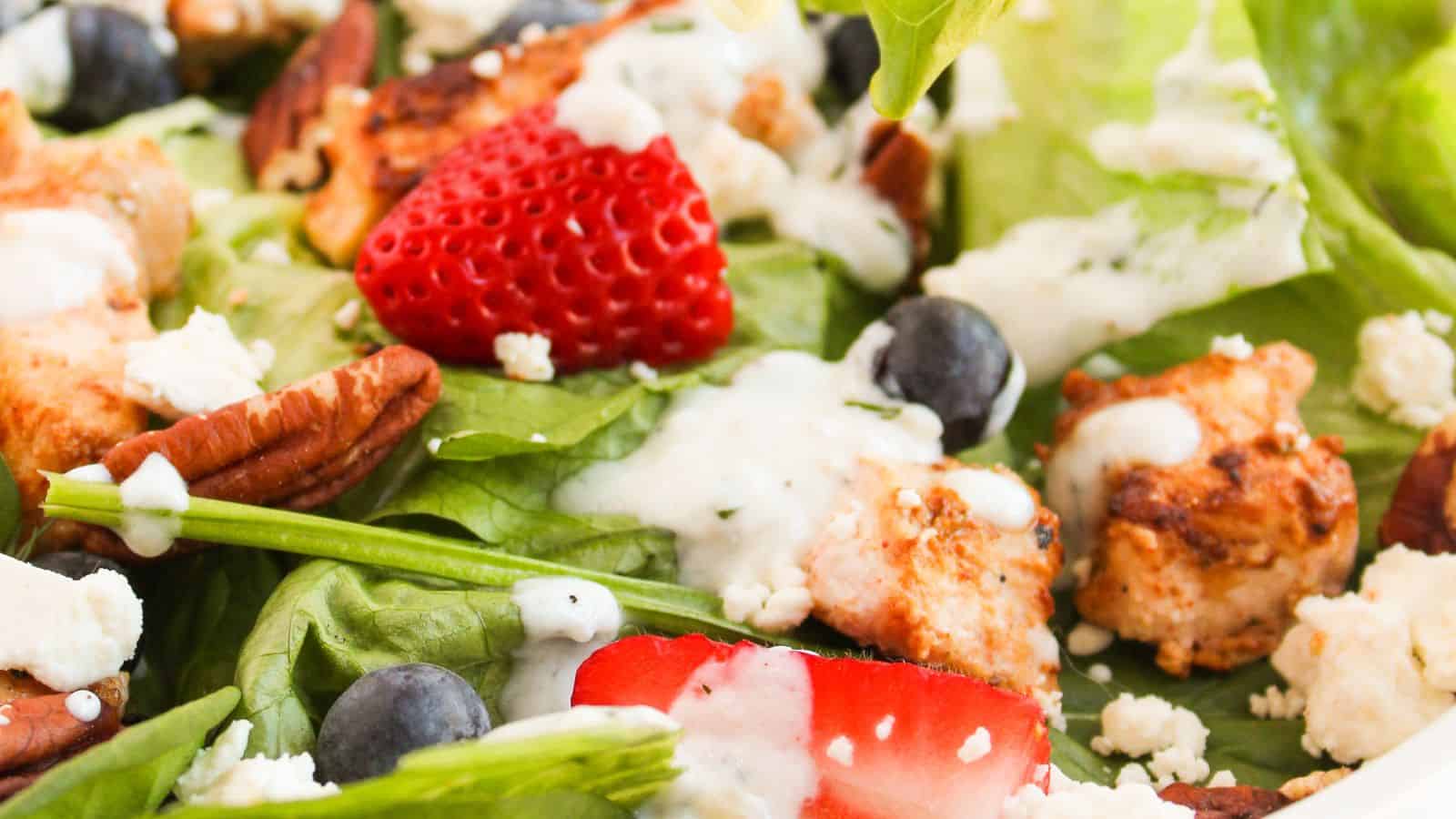 Close-up of a salad with grilled chicken, strawberries, blueberries, pecans, feta cheese, and a creamy dressing on a bed of leafy greens.