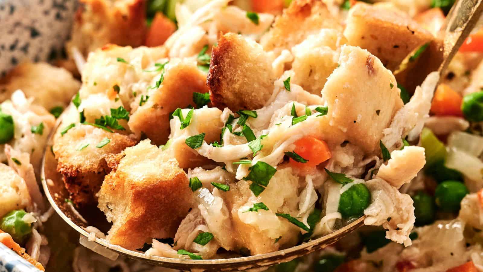 A close-up of a spoonful of chicken stuffing, showcasing croutons, shredded chicken, peas, carrots, and sprinkled herbs.