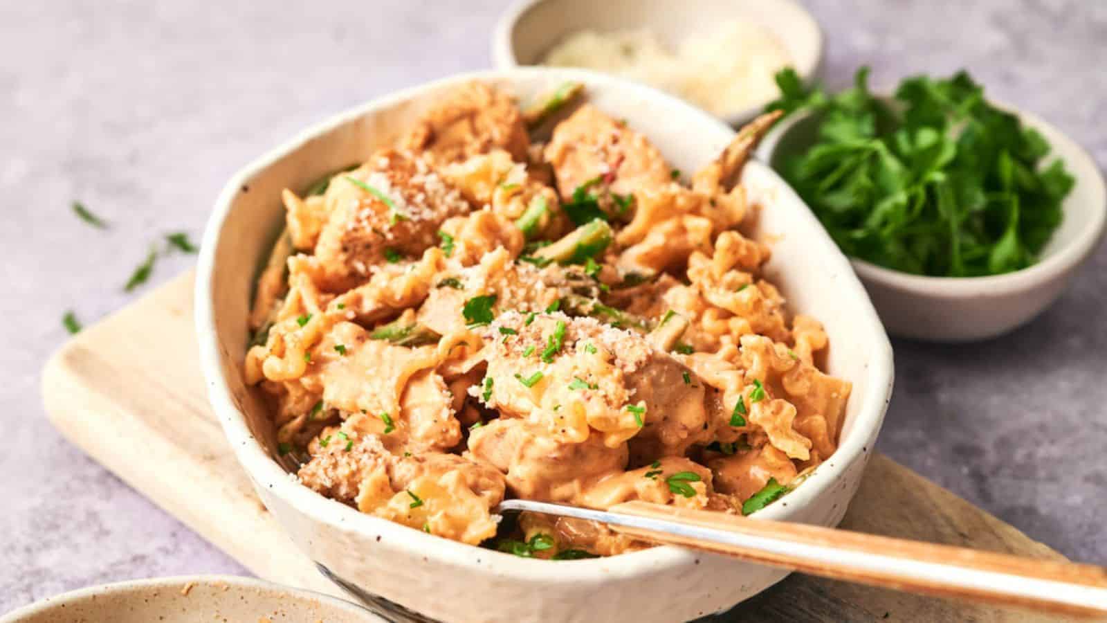 A bowl of creamy chicken pasta garnished with herbs, served with a side of rice and a small bowl of fresh parsley.