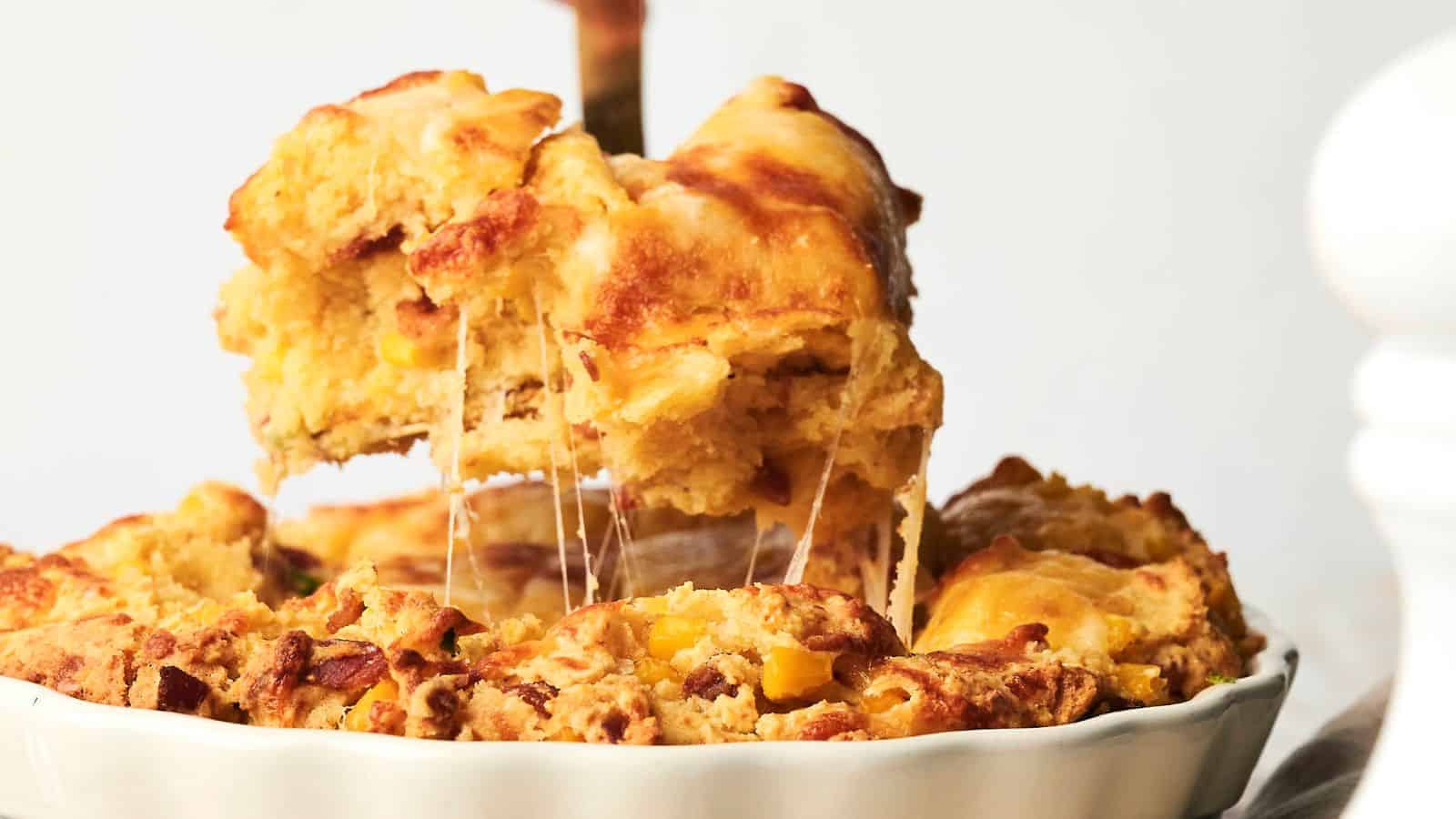 A portion of a cheesy, golden-brown savory cornbread casserole is being lifted from the dish, showing its flaky crust and gooey cheese filling.