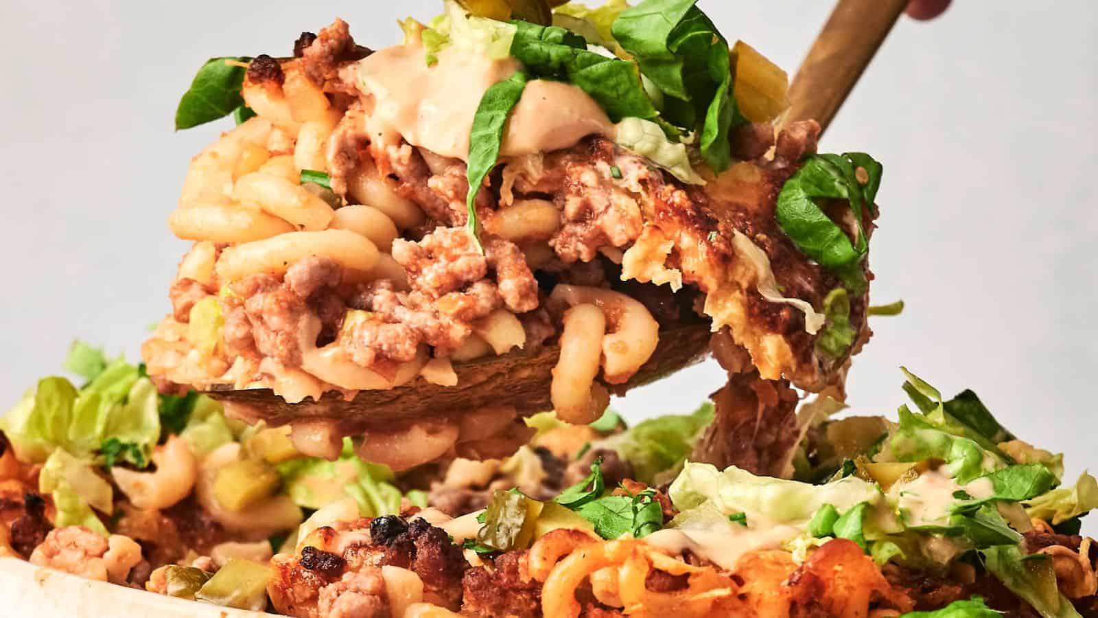 A close-up of a spoon lifting a portion of hamburger casserole, showcasing baked macaroni and cheese layered with ground meat and topped with lettuce and creamy sauce.