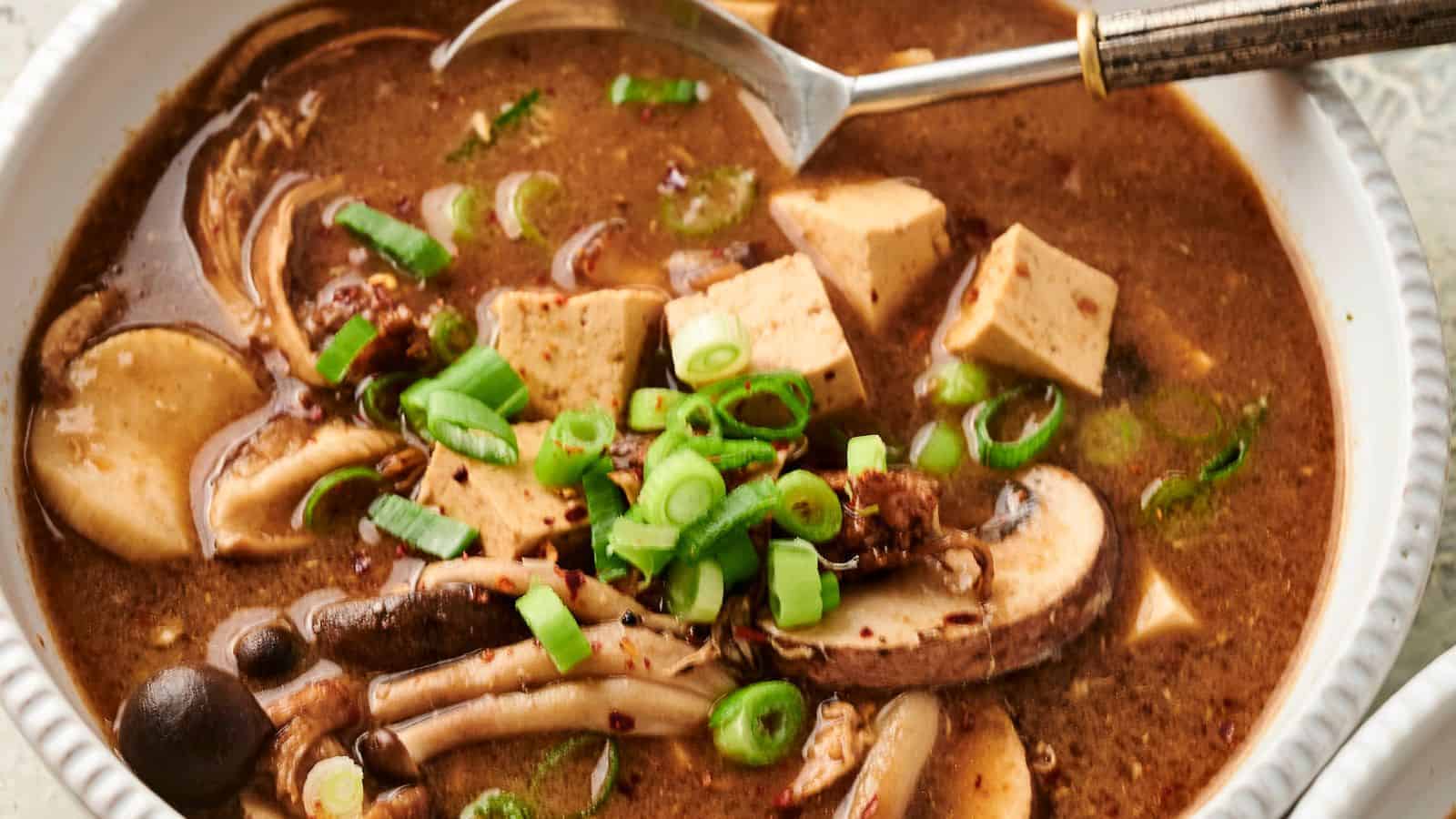 Bowl of hot and sour soup with mushrooms, tofu, and sliced green onions, with a spoon on the side.