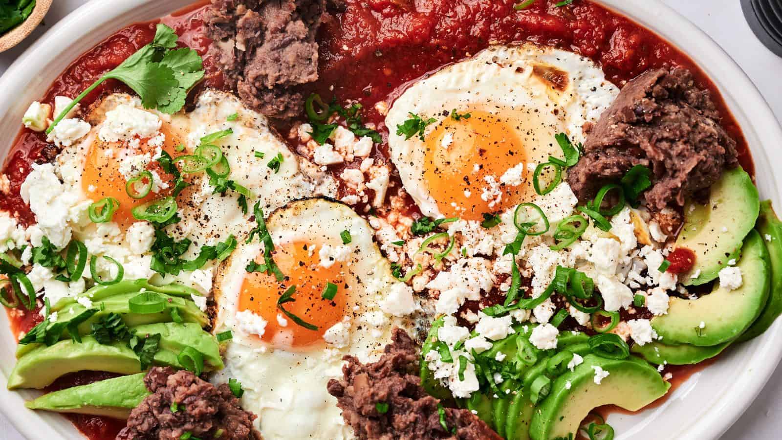 A plate of huevos rancheros with sunny-side-up eggs, avocado slices, refried beans, tomato sauce, crumbled cheese, and garnished with green onions and cilantro.